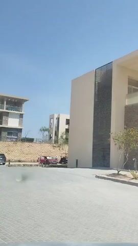 The video presents a slow, panning view of a modern residential or resort complex under a clear blue sky, likely during the daytime. The camera moves from right to left, revealing the architectural style and landscaping of the area.

On the right, a large, beige-colored building with dark-colored vertical accents is seen first. As the camera pans, a paved road made of light-colored interlocking pavers becomes prominent in the foreground. To the left of the initial building, another multi-story building with a light-colored facade, balconies, and stone accents comes into view. This building appears to have multiple units with large windows and glass railings on the balconies.

Between the paved road and the buildings, there are landscaped areas featuring stone retaining walls, sparse trees, and various shrubs, characteristic of a dry climate. A few cars are parked along the side of the road and in designated parking spots. One dark-colored car is visible moving slowly. No people are discernible in the video. The overall impression is that of a well-maintained, contemporary development.