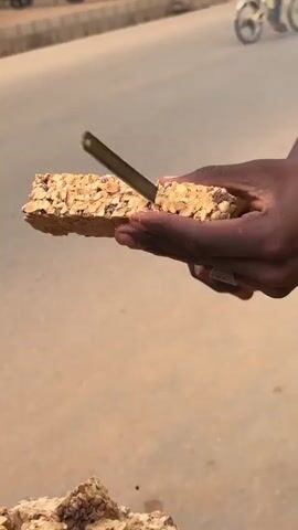 Here's a description of the video:
A street vendor cuts a block of peanut brittle. The person wears a yellow t-shirt. The brittle is cut using a small, dark-handled knife. The cut piece is held in their hands, and crumbs fall as the brittle is handled. The vendor stands next to a pile of similar peanut brittle blocks on a paved surface. In the background, a blurred road and sidewalk are visible.
