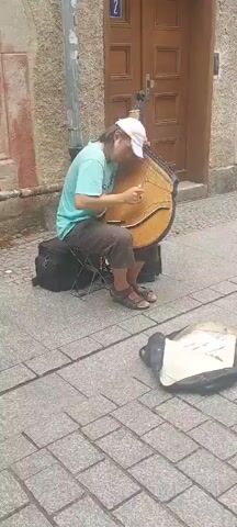 A person wearing a turquoise t-shirt, brown shorts, a white baseball cap, and sandals is sitting on a folding stool and playing a bandura. The bandura is a large, plucked string instrument with a rounded body and numerous strings. The person is seated on a cobblestone sidewalk in front of a building with a brown wooden door. A black bag sits on the ground to the person's left, and a white and black bag is open on the ground in front of them, possibly serving as a collection receptacle. The background consists of a textured wall and the wooden door. The lighting suggests it is daytime.