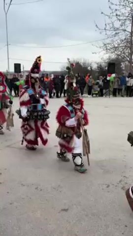 The video shows four people performing a traditional dance outdoors in front of a small crowd. Each dancer is wearing an elaborate, colorful, traditional costume that consists of a fur-trimmed red-and-white robe, patterned leggings and shoes, and a tall, pointed headdress adorned with feathers and other decorations. The costumes also include a series of large bells around the waist, which ring as the dancers move. One of the dancers carries a stuffed animal, another a decorated staff, and a third carries what appears to be a small shovel. The dance seems to involve rhythmic steps and turns, with the bells creating a loud, percussive sound. The crowd in the background includes people of all ages and genders, who are watching the performance with interest. The setting appears to be a village square or a similar public space, with simple buildings and bare trees visible in the background under an overcast sky.
