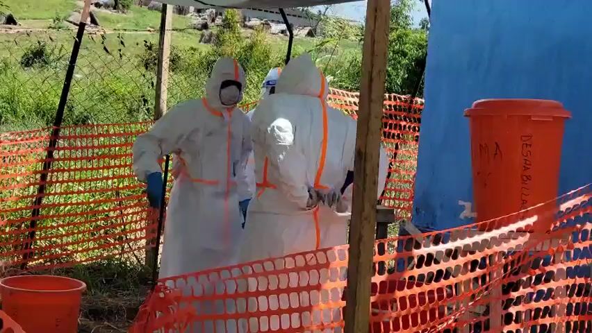 The video captures three individuals dressed in white full-body hazmat suits with prominent orange reflective strips, face masks, and blue gloves. One of the individuals also wears a clear face shield. They are situated outdoors under a makeshift canopy, which is supported by wooden posts and covered with dark and blue tarp-like material.

An orange safety netting acts as a barrier, delineating the operational area. On the ground to the left, two orange buckets are visible, while a larger orange container or drum with partial text including "MA" and "DESHA BILLAR" (or similar characters) is seen on the right.

In the background, beyond a chain-link fence, lush green vegetation, including grass, bushes, and trees, extends into the distance, with natural rock formations or hills visible. The ground within the immediate work area is unpaved dirt. The bright lighting suggests it is daytime.

The individuals appear to be performing tasks or preparing within this designated, cordoned-off zone. The protective gear and setup suggest a public health or emergency response operation, likely a decontamination or screening point, in Tchere, Democratic Republic of the Congo.
