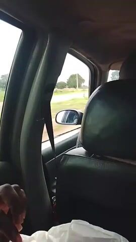 This video captures a view from inside a moving vehicle, likely a car, looking out the passenger side window. The scene is set in a rural or semi-rural area, with a paved road visible in the distance. The surrounding landscape includes green vegetation and scattered trees, with a slightly overcast sky suggesting it might be daytime, possibly morning or afternoon.

Several vehicles are seen on the road, including a truck, a sedan, and a three-wheeled auto-rickshaw (also known as a Keke Napep in Nigeria). The auto-rickshaw is carrying passengers, and in one frame, a group of people are seen gathered on the side of the road, some of whom appear to be interacting with others near a roadside stall or structure.

The interior of the vehicle shows a portion of the seat and a seatbelt. A hand is visible in the lower left corner of the frame, holding a white plastic bag. The overall mood appears to be observational, as if documenting the journey and the passing scenery. There is no visible text within the video.

The context provided indicates the location is Barakin Kuru III, Nigeria, which aligns with the presence of the auto-rickshaw and the general appearance of the landscape. The activity is primarily passive observation of a road trip and the surrounding environment in Nigeria.