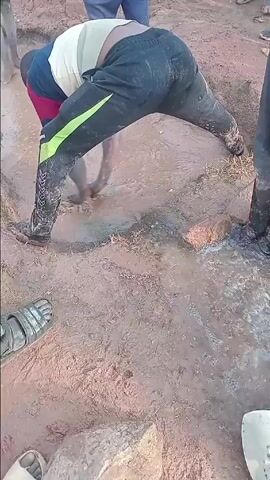 The video, set in Agingi Zion, Nigeria, captures a group of individuals engaged in manual labor in a muddy, outdoor environment.

In the center, a man is bent over, facing away from the camera, actively digging or clearing a muddy pool of water with his hands. He wears a light-colored t-shirt and black sweatpants with a bright green stripe on the side; his pants and lower legs are heavily soiled with mud. Water flows into and through the muddy area where he is working.

In the foreground, another person is kneeling, wearing a grey long-sleeved top and light-colored sandals. This individual is holding a yellow measuring tape, seemingly observing or marking a point on the ground. To the right, another person's legs and feet, clad in dark pants and boots, are visible, standing in a similar muddy area. Additional legs and feet of other individuals can be seen in the background, suggesting a collaborative effort.

The scene takes place on reddish-brown, unpaved soil, characteristic of many regions in Nigeria. Small rocks are scattered across the ground. The presence of significant mud and flowing water suggests either recent rainfall or the active management of a water source. The lighting indicates it is daytime, likely during the late morning or afternoon. The individuals appear focused on their task. No clearly legible text is discernible on clothing or objects, other than indistinct markings on the measuring tape. A silver shovel head is partially visible on the right side of the foreground.