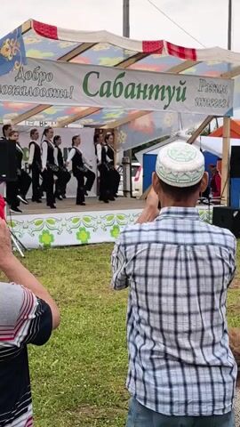A group of young women in matching black vests and white shirts perform a synchronized dance on an outdoor stage. A banner above the stage reads "Добро пожаловать! Сабантуй" which translates to "Welcome! Sabantuy". A man in a plaid shirt and a white and green skullcap films the performance with his phone. Another person's hand is visible in the lower left corner, also appearing to be taking a photo. The stage is decorated with green floral patterns. The ground is covered in grass.