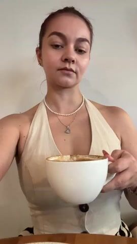 A woman with her hair pulled back in a bun and wearing a pearl necklace and a tan halter top with black buttons is holding a white coffee cup with both hands and drinking from it. Her eyes are looking up and to the left. Her left hand is in the foreground and out of focus. The background is a plain white wall.