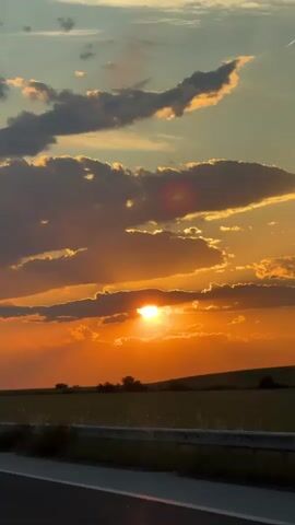 The video shows a colorful sunset landscape from a moving vehicle. The sun is positioned near the horizon, partially obscured by clouds, casting an orange glow on the sky. The landscape includes a green field separated from the road by a gray guardrail. The clouds are a mix of gray and orange. Music plays in the background.