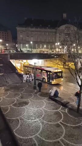 At night, a yellow bus pulls up at an underground bus stop near a city square. Pedestrians board the bus while other people sit on a nearby bench.  The cobblestone square is decorated with a swirling pattern. A grand building stands in the background, illuminated by streetlights. Trees are bare.  A person walks through the square.