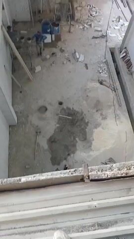 The video shows the interior of a building under construction, filmed from an elevated position. Two construction workers are visible on the ground floor, working near buckets and materials. The floor is mostly concrete, covered in debris and damp patches. A hammer lies on the ground. The camera angle rises to show a partial second floor with unfinished stairs, and then to the corrugated metal roof supported by a metal frame. Another worker is visible on scaffolding near the roof. There is also a cement mixer on the second floor.