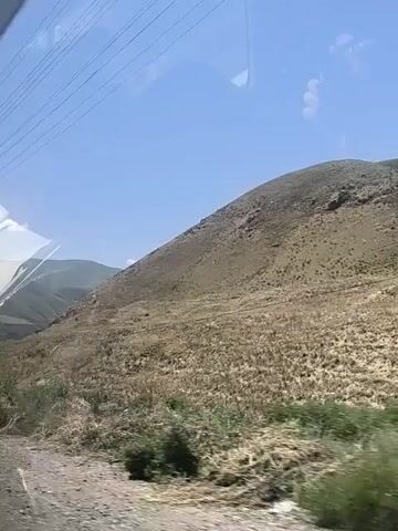 The video shows a view from inside a vehicle, looking out a window at a hilly landscape. The sun is shining and the sky is blue with a few clouds. There are power lines visible across the top of the frame. In the distance, a large, light-colored structure, possibly a dam, is visible on the left side of the frame. The hills are covered in dry grass and sparse vegetation. A wooden utility pole with wires is visible on the right side of the frame. The camera is moving, suggesting the vehicle is in motion.