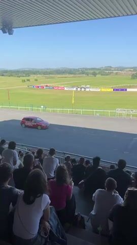 The video shows a race track with a red car driving on it. The car has a circular logo on its side. There are spectators in the foreground sitting in tiered seating. The race track is green with white railings and flags. The sky is blue and there are hills in the background. The video appears to be taken from under a roofed structure.