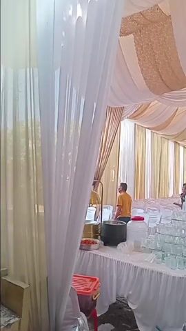 The video captures a lively outdoor event taking place under a large, elaborately decorated tent. The tent's interior is adorned with flowing white and gold or cream-colored fabric drapes, creating a festive and elegant atmosphere. Some drapes also hang vertically along the sides, resembling curtains. A chandelier is visible hanging from the draped ceiling.

At the beginning of the video, the camera focuses on a section of the tent where food and drink stations are set up. White-skirted tables hold several large silver chafing dishes, a big black pot, and numerous clear plastic cups stacked in pyramids. Several men are gathered around these tables; one in a yellow t-shirt is attending to the food, while others observe or converse. A woman in a blue hijab is also seen near the food station, and another woman in a patterned dress approaches the table.

As the camera pans, it reveals rows of neatly arranged white chairs, each adorned with a blue sash, indicating a formal event like a wedding reception. Many guests are already seated, facing a beautifully decorated stage or altar at the far end of the tent, which features floral arrangements. The ground under the tent appears to be dirt or gravel.

The camera then pans towards the tent's exit, showing a glimpse of the outdoor surroundings with trees and another covered area in the distance. The sun is shining brightly, suggesting a clear day. A woman in a pink hijab walks through this area.

Returning to the main tent, the camera again shows the food and drink area where more people are congregating. A man wearing an orange t-shirt with "BERASTA" printed on the back is seen near the drink dispensers, which include large clear water containers and stacks of clear cups. Boxes with an "AQ" logo are also visible, likely containing bottled water. Throughout the video, people are seen mingling, serving themselves, and interacting, giving the impression of a joyous community gathering. The overall ambiance is bright, airy, and celebratory.
