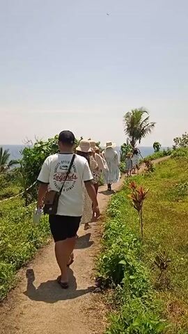 A group of people walks along a paved path towards the sea on a sunny day. A man in a white t-shirt, black shorts, a black cap, and brown sandals leads the group. He has a brown crossbody bag and carries a blue water bottle. The group behind him consists mainly of women in light-colored dresses and wide-brimmed hats. The path is flanked by green grass and bushes. A lone palm tree stands to the right of the path. The blue ocean is visible in the distance against the clear sky.