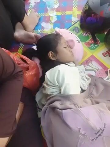 The video begins with a close-up of a young girl sleeping on a colorful mat, covered by a light purple blanket. She has dark hair and is wearing a light-colored shirt. Next to her is a pink stuffed animal. The camera then pans to a woman wearing a hijab, who appears to be taking a selfie. She is joined by a man next to her, and they are both sitting under a striped awning. The woman leans her head on the man's shoulder, and they both look towards the camera. The background suggests they are at an outdoor venue, possibly an amusement park, with a Ferris wheel structure visible in the distance. The video appears to capture candid moments of rest and affection between the individuals.