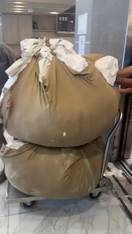 Two men are pushing a two-tiered cart laden with large, tan laundry bags. The cart has a metal frame and wheels. The top laundry bag is overflowing with white and off-white fabric. The bottom laundry bag appears to have a greenish discoloration. One man, wearing a dark blue short-sleeved shirt and brown shorts, is pushing the cart from behind. The other man, wearing a light blue short-sleeved shirt, is on the left side of the cart, helping to push. In the background, a reception desk is visible with a person sitting behind it. A sign with red lettering that reads "B2" is also visible on the wall. The floor is a marble-like pattern.