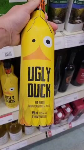 A hand holds a bottle of "Ugly Duck" wine, which is designed to look like a yellow duck face with large, googly eyes and an orange beak. The bottle has a red label with the words "UGLY DUCK" in bold black lettering. Below that, it says "RED BLEND CABERNET SAUVIGNON MERLOT" and "750 mL 13.5% alc/vol RED WINE/VIN ROUGE". The bottle is on a shelf in a store, with other bottles and boxes visible in the background. The hand is holding the bottle from the neck, and the fingers are visible. The background is slightly blurred, but it appears to be a wine or liquor store aisle.