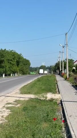 A vertical video shows a rural road on a sunny day. The road curves to the left, with a paved sidewalk and grassy embankment on the right. Trees line the left side of the road, and more trees and hills are visible in the distance, topped with two wind turbines. Two white vans and a black car are driving away from the camera on the road. The sidewalk is lined with streetlights and small red flowers. The sky is clear and blue.