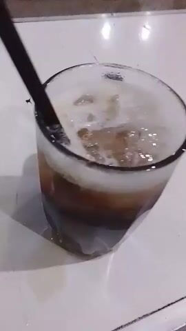 The video shows a close-up, slightly shaky, view of a glass filled with a dark liquid and ice. A black straw is visible in the glass. The glass is sitting on a white surface, possibly a table, with some condensation or liquid visible around the base of the glass. The background is out of focus, but appears to be an indoor or semi-outdoor setting with some indistinct shapes that could be furniture or walls. The lighting is dim, creating shadows.