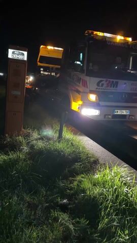 A tow truck, with the words "GARAGE FLOURY" and "G3M AUTO" prominently displayed, is parked on the side of a road at night. Its headlights are on, illuminating the roadside. The scene includes a roadside marker with the number "A31 243+000" visible. The truck has flashing lights on its roof, indicating it is there to assist a stranded vehicle. Some grass and road barriers are visible. The overall setting suggests a roadside assistance situation at night.