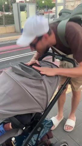 A man in a brown t-shirt, beige shorts, white slides, and a white baseball cap is pushing a grey stroller on a sidewalk. He has a backpack on his shoulders and a small beige bag slung across his chest. He leans down to check on the baby in the stroller, smiling. The stroller has a grey canopy and some items are visible inside. In the background, there is a sidewalk with pink lines, a fence, some trees, and a modern-looking building with glass doors. The lighting suggests it is daytime.