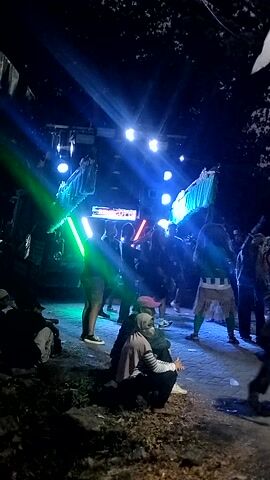 The video shows a nighttime outdoor event with a stage and lighting. Several people are visible, some dancing or interacting near the stage, while others are further back. There's a flag visible, held by two individuals, which appears to be red and white. The stage is illuminated with bright blue and green lights, and there's a sign with text on it. The ground is uneven, with some areas appearing to be dirt or gravel. The overall atmosphere suggests a festive or musical gathering.