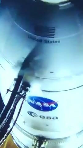 The video begins with a blurry shot of what appears to be a person in an orange flight suit inside a cockpit, with a shiny black helmet in the foreground. The camera then pans to a close-up of the helmet, showing reflections of lights. Following this, the scene shifts to a rocket launch. The rocket, a white cylindrical object with black markings, is seen ascending from a launchpad amidst a plume of white smoke. The launchpad structure is a dark, metallic framework. The background is a vibrant green, suggesting an outdoor setting, possibly during daytime. The overall event is the launch of a rocket. There is no discernible text visible in the video. The location context of Homyel, Belarus, does not provide any direct visual cues within the provided frames.