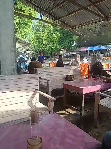 The video shows an outdoor covered seating area with several tables and benches. People are seated at some of the tables, and some are standing. The environment appears to be a casual dining or gathering spot, possibly near a body of water as indicated by the reflections and the presence of what looks like a dock or boats in the background. The lighting suggests it is daytime.