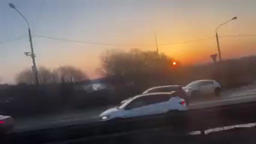 The video shows a view from a car driving on a highway, likely an overpass or elevated road. Several other cars are traveling on the highway in the same direction. In the background, a neighborhood is visible, partly covered in snow. The sun is setting, creating a vibrant orange and yellow sky. There are trees and utility poles along the roadside. The video appears to be taken at either sunrise or sunset, given the low angle of the sun.
