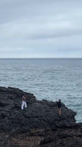 The video shows two people on a rocky, dark-colored coastline with the ocean stretching to the horizon under a cloudy sky. One person is wearing white pants and a brown top, while the other is wearing a dark outfit and is barefoot. The person in white walks across the rocks while the other one appears to be taking photos or recording video. The waves are visible lapping against the rocks.