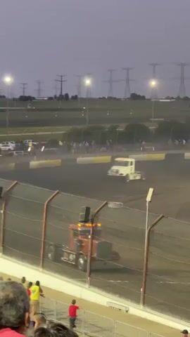 The video shows a nighttime truck race on a dirt track. A white truck with flashing red lights is in the foreground, kicking up dirt as it turns. In the background, two orange trucks are also racing. Spectators are visible in the lower portion of the frame, behind a chain-link fence. Power lines and lights are visible in the distance. The sky is dark and appears to be overcast.
