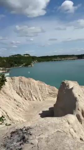 The video shows a view of a lake and beach from a high vantage point. The sky is blue with white clouds. The lake is a clear turquoise color. The beach is small and sandy, and several people are sunbathing, swimming, and relaxing. The landscape in the foreground consists of white rock formations with deep grooves. There are signs of a previous fire on one of the rocks. A boat is visible on the lake, further offshore. In the distance, there are treelines along the shoreline. The camera slowly pans to the right, showing more of the beach and the people enjoying the scenery. Music plays in the background.