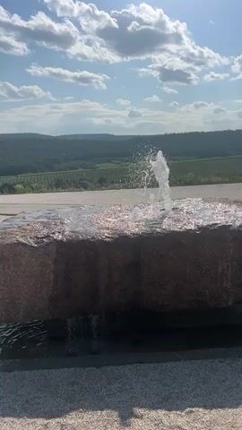 A close-up shot focuses on a small stone fountain with water spraying upwards from its surface. The stone is rough and reddish-brown. In the background, a rolling landscape of green hills covered in trees and vineyards stretches out under a bright blue sky with fluffy white clouds. The sun is visible in the upper right corner of the frame. The video captures the movement of the water and the natural beauty of the surrounding environment.