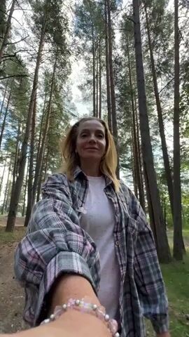 A woman with blonde hair and a pink and gray plaid shirt is walking in a forest. She is smiling and looking to the side. The camera is following her from behind and is also holding a bracelet with pink beads. The path is dirt and surrounded by tall pine trees and green foliage. The sun is shining through the trees, creating dappled light on the ground. The woman is walking away from the camera and then turns to face it briefly. The video then shows the path ahead of her, disappearing into the trees.