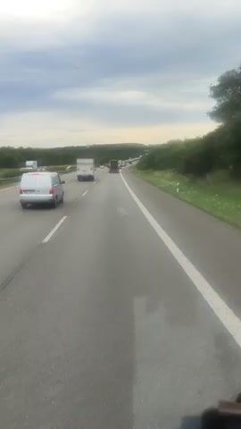 A POV shot from a vehicle traveling on a highway shows traffic ahead. The road is asphalt with white lane markings. On the left lane, a white van is visible. In the right lane ahead, several trucks and cars are driving. The surrounding environment consists of green trees and vegetation on both sides of the highway, and the sky is overcast with clouds. The video is a continuous shot with no apparent significant changes in the scene.
