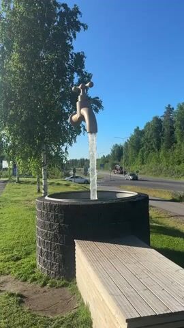 The video shows a large faucet sculpture, appearing to pour water into a circular stone basin. The sculpture is located outdoors, with trees and a road in the background. The sun is shining, and the sky is clear blue. There are a few cars driving on the road in the distance, but no people are visible in the immediate vicinity of the sculpture. The water appears to be real, creating a continuous stream that falls into the basin, which has a dark, rough texture. Surrounding the basin is a wooden bench or platform. The overall scene is serene and picturesque, showcasing an unusual piece of public art.