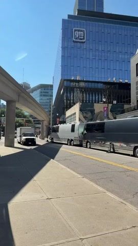 The video shows a street scene with several large, dark-colored buses parked on the right side of the road. In the background, a modern glass building with a "GM" logo is visible, along with other buildings and an elevated train track. A white truck is also visible on the left side of the street, near the train track. The sky is clear and blue, indicating a sunny day. The camera pans slightly to the right, focusing on the buses and the buildings. There are no people clearly visible in the video.