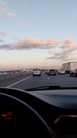 A view from inside a car driving on a highway.  Several vehicles are in the frame including cars, vans and a semi-truck.  The sky is visible with pink clouds and the moon. Music is audible in the background.