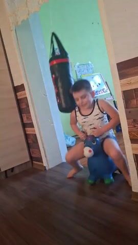 A child bounces on a blue inflatable toy, then falls off and lands on the floor. The child is wearing a white tank top with black stripes and dark blue shorts. In the background, a black punching bag hangs from the ceiling, and a shelf with toys is visible. The room has wooden floors and pale green walls.