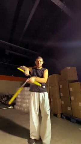 The video shows a man in a sleeveless black shirt and loose white pants juggling two yellow objects that appear to be nunchucks or similar practice weapons. He is standing in a dimly lit indoor space that resembles a warehouse or storage area. Stacks of boxes and wrapped pallets are visible in the background, along with exposed ceiling pipes and lighting fixtures. The man is in the process of performing a juggling routine, with the objects in motion.
