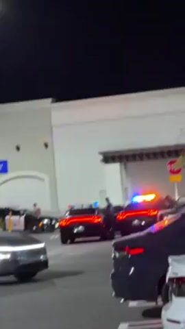The video depicts a nighttime scene in front of a building with a "STOP" sign visible. Several police cars, with their emergency lights flashing in blue and red, are parked in what appears to be a parking lot. Police officers are seen standing near the vehicles. There are other cars parked nearby, and the overall impression is that something is happening that requires police presence. A man is heard saying "We're at the hallmark down the street, that's where the action's happening."