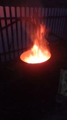 A close-up shot focuses on a metal barrel with a bright orange fire burning inside. The flames rise and flicker, with sparks occasionally flying upwards. Behind the barrel, a white picket fence is visible, silhouetted against the dark background. The scene appears to be outdoors at night, with the fire illuminating the immediate surroundings. The bottom portion of the frame shows dark ground, possibly soil or grass. To the right of the barrel, a piece of white material, possibly cardboard or paper, is lying on the ground. The video is static with no movement captured.