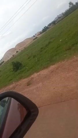 The video shows a view from inside a vehicle, looking out the side mirror. The vehicle is traveling on a dirt road. To the left of the road is a grassy field. In the background, there are hills and a cloudy sky. A power pole is visible in the distance. The vehicle passes an abandoned building with a partially damaged roof and graffiti on its walls. Later, two people on a motorcycle pass by the vehicle. The person in the back appears to be wearing a beanie. The video is filmed from a moving vehicle.