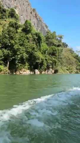 The video shows a body of water from the perspective of a moving boat. The water is greenish-grey and churning behind the boat. The shoreline is covered in lush green trees and vegetation, which extends up the side of a large, rocky cliff face. The sky is clear and blue.