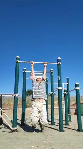 A man performs a gymnastic routine on an outdoor pull-up bar. He starts by hanging from the bar, then pulls himself up, tucks his knees, and flips upside down, his legs going over the bar. He then brings his legs down and over the bar again, ending in a seated position on the bar. The setting is an outdoor gym with a clear blue sky and a patch of dry grass in the background. The man is wearing a striped t-shirt, light-colored shorts, and a hat.