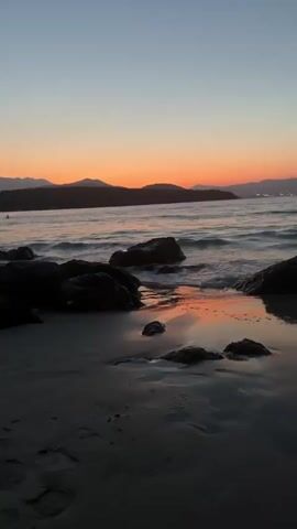 This is a video of a beach at sunset. The water is calm and waves are gently lapping at the shore. There are several dark rocks scattered on the beach and in the water. In the distance, mountains are silhouetted against the orange and pink sky. The sky transitions from a deep blue at the top to a vibrant orange and pink near the horizon. The overall mood is peaceful and serene, evoking a sense of tranquility. The time of day is clearly sunset due to the colors of the sky. There are no visible objects or text in the video. The location is a beach, as indicated by the sand, rocks, and sea.