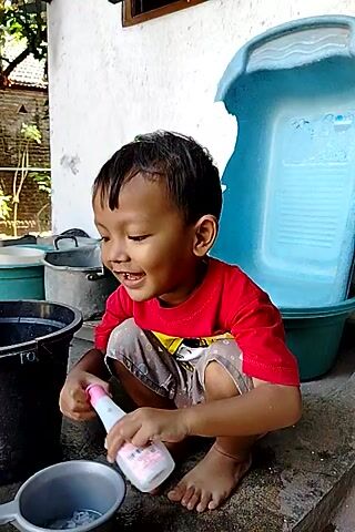 A young boy wearing a red t-shirt and patterned shorts is crouching on the ground. He is smiling and looking down at a white bottle with pink accents that he is holding in his right hand. His left hand appears to be holding a black ladle with a white handle. He is near a large, light blue plastic tub and several other dark-colored containers. The ground is made of concrete and dirt. The background shows a brick wall and some vegetation. The video is oriented vertically.