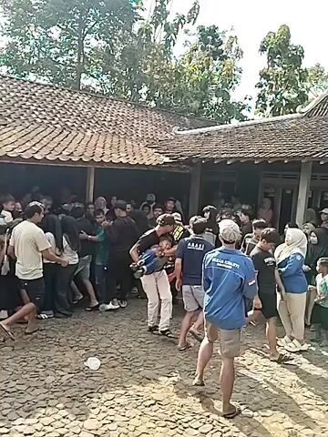 A crowd of people is gathered outside a building with a tiled roof. The ground is covered in gravel and dirt. In the foreground, a man wearing a black shirt and white pants is holding a young child. To his right, another man in a blue shirt with white writing on the back is standing. The crowd consists of men, women, and children of various ages. Some individuals are looking towards the building, while others appear to be interacting with each other. The overall impression is of a busy outdoor gathering.