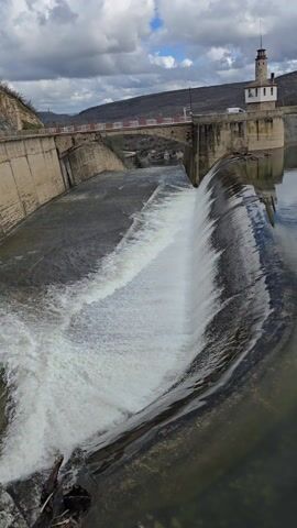 The video shows a dam with water flowing over its edge, creating a waterfall effect. The dam appears to be made of concrete and has a bridge spanning across it. A small tower-like structure stands on the side of the dam, adding to the architectural landscape. The sky is overcast with dark clouds, suggesting a potentially gloomy day. The surrounding environment seems hilly and sparsely vegetated. The camera angle is positioned at an elevated perspective, providing a comprehensive view of the dam, water flow, and the surrounding landscape.