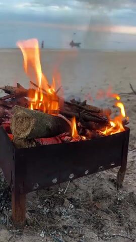A close-up shot of a campfire burning on a sandy beach. The fire is in a rusted metal grill with wooden legs. Logs and sticks are burning, with flames reaching high. In the background, a calm body of water is visible, with two figures standing on the shore and a small boat in the distance. The sky is overcast with muted colors. The overall impression is one of a quiet, serene evening by the water.