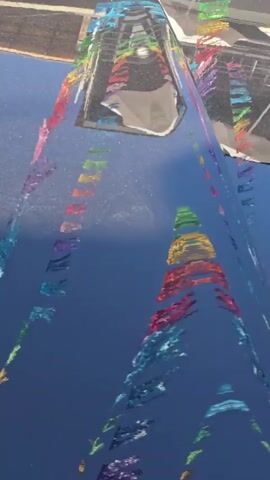 The video shows a close-up, abstract view of a metallic, reflective surface, possibly a car hood or a large sculpture. The surface is reflecting an outdoor environment with a bright blue sky and a structure that appears to be a building or a bridge. What makes the video unique is the presence of a string of colorful, fragmented flags or pennants draped across the reflective surface. These colorful elements are distorted and stretched by the curves and reflections of the metallic surface, creating a mesmerizing, kaleidoscopic effect. The colors of the flags range from red, orange, yellow, green, blue, and purple, arranged in a repeating pattern. The camera appears to be slowly moving, causing the reflections and the colorful fragments to shift and shimmer. There are no visible people or clear actions taking place, beyond the implied movement of the camera.