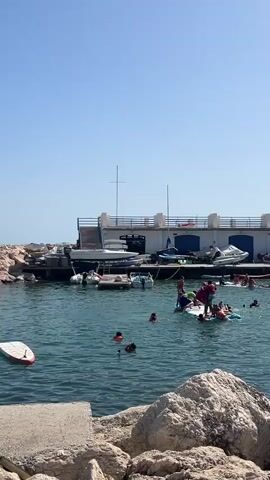 The video depicts a lively scene at a marina under a clear blue sky, suggesting a sunny day. The water is a deep blue, with several individuals engaged in various water activities. In the foreground, a red and white paddleboard bobs on the water, and large rocks line the shore.

The main activity centers around a group of people on and around paddleboards. One person, wearing a pink top and dark shorts, appears to be instructing or assisting others on a larger, green inflatable watercraft. Several other individuals, some wearing red life vests, are in the water, swimming or paddling. One person is captured mid-splash, suggesting an energetic or playful moment.

In the background, a building with a white facade and blue accents sits on a dock. Several boats are moored here, including a sleek white motorboat docked above the water. Smaller motorboats are also visible. The overall impression is of a recreational area where people are enjoying a warm day by the sea, participating in water sports and swimming. The atmosphere appears cheerful and active.